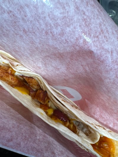 The image shows two soft tacos inside a paper wrapper. The tacos appear to contain ingredients such as beans, shredded cheese, and pieces of cooked chicken or meat. The focus is on the food, with the paper wrapper acting as a backdrop. The image does not contain discernible information relating to the city of Saldus, Latvia, or people, actions, interactions, or any further details about the environment.