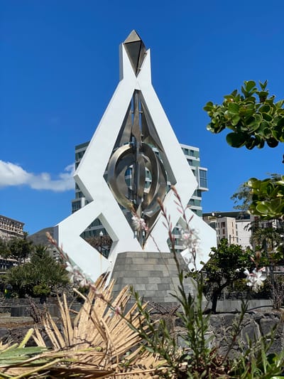 The image depicts the "Plaza de España" monument in Santa Cruz de Tenerife, Spain. The monument is a prominent white structure with geometric shapes and a central metal sculpture composed of concentric circles. In the background, buildings and the sky are visible, with a few scattered clouds. In the foreground, there is vegetation, including trees, bushes, and flowers. The image appears to be taken from a slightly low angle, looking upwards towards the monument. The lighting suggests a sunny day, with clear blue skies.