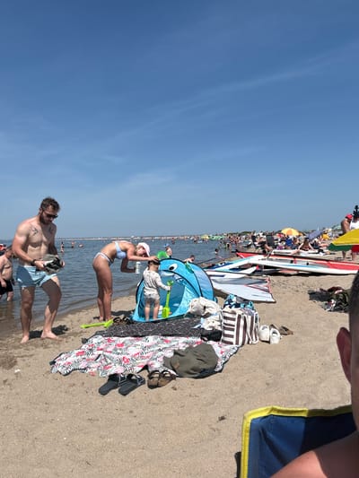 On a sunny day at the beach in Rewa, Poland, a group of people are enjoying the water and sand. In the foreground, a man in light blue swim trunks is holding a dark towel, looking down at it. Beside him, a woman in a light blue bikini is bending down to attend to a young child inside a blue pop-up tent. The child is wearing light-colored clothing and holding a yellow shovel. A colorful beach blanket with a floral pattern is spread out on the sand behind them, with several pairs of footwear and bags placed on and around it. Further in the background, numerous people are in the shallow water, some swimming and others standing. Several windsurfing boards are propped up on the sand, suggesting a popular spot for water sports. The sky is clear and blue, indicating good weather conditions for a day at the beach.
