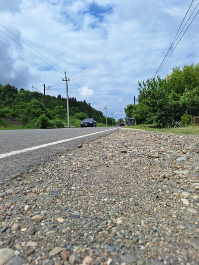 The image depicts a low-angle view of a paved road with a gravel shoulder, suggesting it is in a rural or semi-rural area. Power lines run parallel to the road, supported by utility poles. Lush green trees and vegetation line the side of the road, with a hill covered in trees in the background. The sky is partly cloudy with a mix of blue sky and white clouds. On the road, a dark-colored car is visible in the distance, and further down, a truck and what appears to be a large grey trash bin are present. The scene appears to be located in the region of ნიგვზიანის თემი, Georgia, given the context provided.