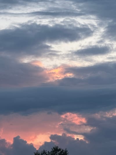 The image shows a cloudy sky with muted colors, suggestive of twilight or an overcast day. The clouds are layered, with some appearing dark gray and others lighter, tinged with hints of pink and orange where light is breaking through. The overall impression is one of atmospheric depth and diffused lighting. There are no people, specific landmarks, or any other discernible objects that would indicate a specific location within a city.