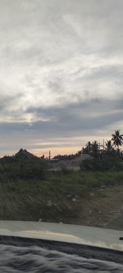 This is a landscape photo taken in Glokaron, Indonesia. The scene depicts a slightly overcast sky with layers of grey and white clouds, suggesting either early morning or late afternoon. In the middle ground, there is a prominent pile of grey sand or gravel, and behind it, a tall, narrow smokestack from an industrial building. To the right of the sand pile, a cluster of palm trees stands out against the sky. The foreground is filled with lush green vegetation, including grass and bushes, with some scattered debris visible on the ground. The bottom of the image shows the reflective surface of what appears to be a vehicle's hood or roof, with a distorted reflection of the sky and trees. The overall impression is one of a rural or semi-industrial area at dusk or dawn, with a hint of industrial activity in the distance. There is no visible text in the image.