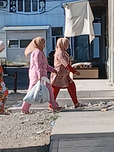 The image shows two women walking on a sidewalk in front of a building in Gulistan, Uzbekistan. The woman on the left is wearing a pink hijab and a pink robe over red pants, carrying a white plastic bag. The woman on the right is wearing a patterned hijab and a patterned robe over red pants. The building behind them has windows and a white sheet hanging from an overhang. The ground is paved with stones and debris.