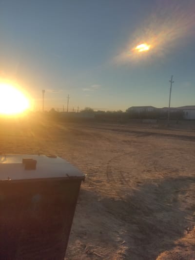 A low-angle, wide shot captures a dusty, outdoor scene during what appears to be sunset or sunrise in Aral, Kazakhstan. The sun, a bright, intensely luminous orb, dominates the left side of the frame, casting long, warm rays across the sandy, uneven ground. To the right, a halo effect surrounds a patch of hazy sunlight in the sky, tinged with orange and yellow hues.
In the foreground, the dark, rectangular structure of a dumpster is partially visible on the left. Its lid is flat and appears to be made of a lighter material, possibly metal. The ground is a light brown, dry dirt or sand, with visible tire tracks etched into its surface. A scattering of small debris and footprints is also present.
In the midground and background, the landscape is sparsely populated with a few buildings and structures. A tall, slender radio tower stands prominently behind the dumpster. Further back, a line of trees or bushes creates a soft, blurred silhouette against the horizon. Several utility poles with wires are scattered across the scene, typical of a rural or semi-urban environment. In the distance, the outlines of buildings, possibly houses or low-rise structures, are visible, some appearing lighter in color. The sky transitions from a pale blue near the horizon to a deeper hue overhead. The overall impression is one of a dry, arid climate bathed in the warm glow of a low sun.