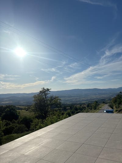 The image depicts a landscape in the city of Tserovo, Bulgaria, featuring a blue car on a road adjacent to lush greenery and a mountain range in the background. Overhead power lines stretch across a bright blue sky with scattered clouds. To the right of the frame is a concrete wall with square tile patterns. The sun is shining brightly, positioned near the bottom left corner.