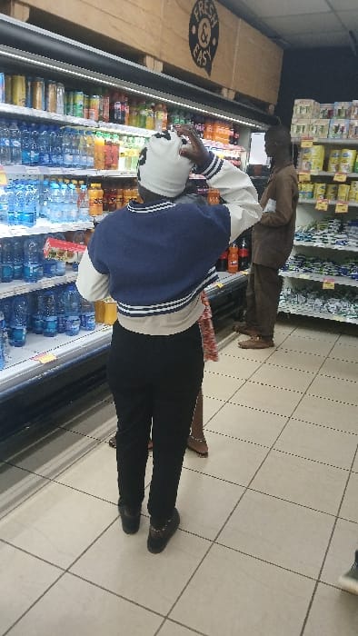 A person wearing a white hat, white and blue striped jacket, and black pants stands in front of a refrigerator case filled with drinks at a grocery store. The person appears to be reaching inside the case, possibly to retrieve an item. Another person wearing a brown suit stands behind them near the top row of shelves with groceries. The tiled floor and the "Fresh N Easy" sign on the left side suggest the setting is a supermarket.