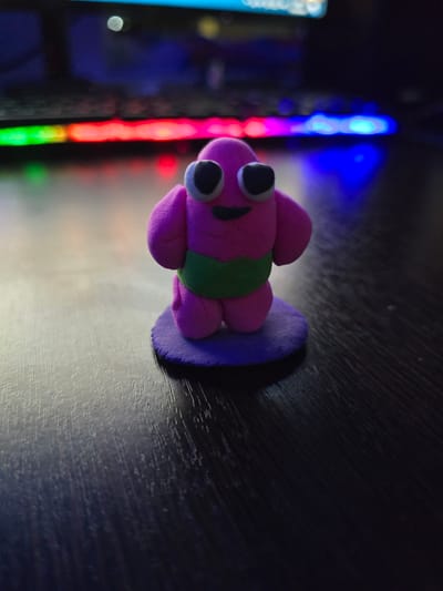 A small, handmade pink clay figure with large white-rimmed black eyes and a black smile stands on a purple circular base. The figure has stubby arms and legs and is wearing a green band around its waist. It is positioned on a dark, textured wooden surface. In the background, out of focus, are the colorful lights of a keyboard, suggesting the figure is placed in front of a computer setup. The lighting is dim, with the colorful keyboard lights creating reflections on the surface. The location within the city of Shali, Russia, cannot be determined from the provided image.