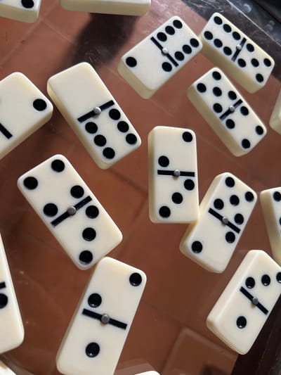 The image shows a close-up, overhead view of several white dominoes scattered on a reflective brown surface. The dominoes are arranged in a non-uniform pattern across the frame. Each domino is divided by a black line and has black dots (pips) on each half, indicating different numerical values. The lighting creates highlights and shadows on the dominoes and the surface, suggesting an indoor environment. No people or specific locations within Villa de Cura, Venezuela, are discernible from the image. The focus is solely on the arrangement and appearance of the dominoes.