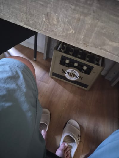 The image shows a person's legs and feet, wearing light gray shorts and beige slides. The person is sitting under a wooden table. On the floor, to the right of the person's legs, is a case of Warsteiner beer bottles. The environment appears to be indoors, with a wooden floor and table legs visible. The image does not provide any information about the city of Offenburg, Germany.