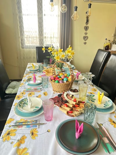 A dining table is set for Easter, featuring patterned plates, glasses, and folded napkins shaped like bunny ears. A basket filled with colorful Easter eggs and daffodils serves as a centerpiece, with a platter of sandwiches, cheese, and chocolate nearby. The scene is set indoors, with chairs around the table and a window visible in the background. The location is Bryastovets, Bulgaria. No people are directly visible in the frame.