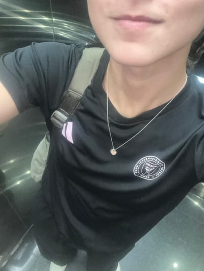The image displays a partial, close-up view of an individual, from the chin to the upper thighs, captured in a selfie within an indoor, highly reflective environment, consistent with an elevator or modern building interior, located in Santa Catarina, Mexico. The subject is wearing a black short-sleeved t-shirt featuring two light pink Adidas stripes on the left shoulder and a white circular "CLUB INTERNACIONAL DE FÚTBOL MIAMI MMXX" crest on the right chest. A thin silver-colored chain necklace with a small, light-colored heart-shaped pendant hangs around the neck. A grey or olive-green backpack strap is visible over the left shoulder, with an attached light-colored fabric item, possibly part of the backpack or another bag, behind the arm. Dark pants or leggings and light-colored footwear are partially visible at the bottom of the frame. The background consists of dark, reflective surfaces exhibiting horizontal light streaks. The individual's chin and mouth are visible, with the lips naturally closed. No other persons or significant actions are depicted.
