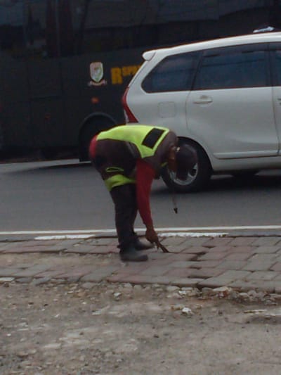 In Megamendung, Indonesia, a person wearing a bright yellow vest and red pants is bent over, using a tool to work on a brick-paved sidewalk. A white SUV is parked in the background, next to a dark vehicle with a logo and lettering that reads "RSP". The ground is a mix of pavement, dirt, and debris, indicating a street or roadside setting.