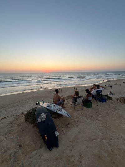 A beach scene at sunset. Several people are sitting in chairs by the cliff. Two surfboards lay on the ground next to a person. A few people walk along the shoreline. The ocean meets the sky, with the sun setting on the horizon.