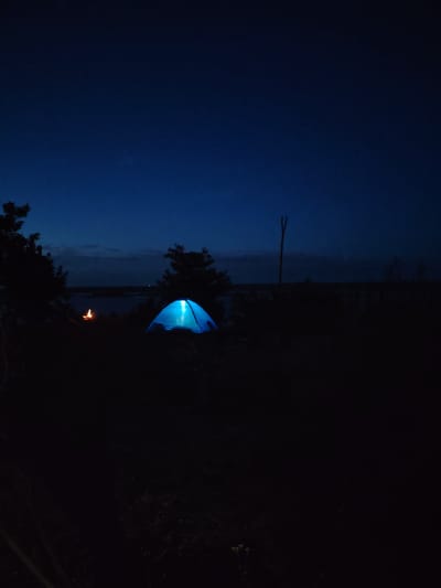 An illuminated blue tent is set up in a dark, natural environment, possibly Staiky, Ukraine, at dusk or night. A small campfire flickers to the left of the tent. Silhouetted trees frame the scene, with a body of water and a faint horizon visible in the background. The sky is a deep blue, transitioning to black at the top of the frame, with a few faint stars. A vertical wooden post stands in the middle ground. The overall impression is one of a remote camping or outdoor setting.
