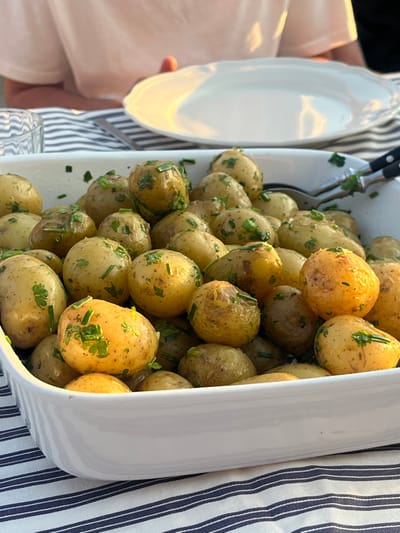 The image presents a close-up, slightly elevated view of a white rectangular ceramic serving dish filled with numerous small, cooked, golden-skinned potatoes. The potatoes are liberally garnished with finely chopped green herbs, consistent with a mix of parsley and chives. A metallic serving spoon and a partial view of a matching fork, both with black handles, are inserted into the potatoes on the right side of the dish.

The dish rests on a blue and white striped tablecloth. In the upper background, an empty white dinner plate with a fluted rim is visible to the right, bathed in warm, natural light. To the upper left, a segment of a clear glass object is present. Directly behind the table setting, the upper torso, right arm, and right hand of an individual wearing a light-colored short-sleeved top are partially visible. While the specific location is indicated as Skagen, Denmark, no visual elements within the image confirm this geographical detail.