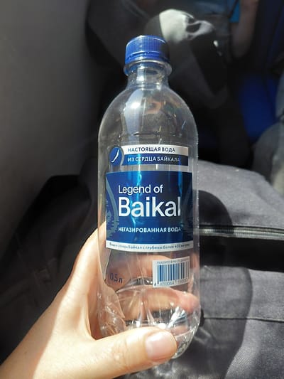A clear plastic bottle of "Legend of Baikal" water is held in a person's left hand. The bottle has a blue cap and a blue and white label. The label prominently displays "Legend of Baikal" in large white letters. Smaller text on the label indicates "НАСТОЯЩАЯ ВОДА ИЗ СЕРДЦА БАЙКАЛА" (True water from the heart of Baikal) and "НЕГАЗИРОВАННАЯ ВОДА" (Still water). Below this, it states "Вода из озера Байкал с глубины более 400 метров" (Water from Lake Baikal from a depth of over 400 meters). A barcode is visible on the lower part of the bottle. The bottle appears to be about half full. The background is dark and out of focus, suggesting an indoor environment or a shaded outdoor area. The lighting suggests it is daytime. The location is not explicitly stated in the image but the brand "Legend of Baikal" refers to Lake Baikal, which is located in Siberia, Russia. The city of Chulym, Russia is geographically located in the Novosibirsk Oblast, which is part of Siberia, making it a plausible general location within the broader region associated with Lake Baikal.