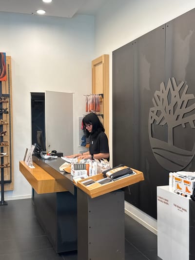 The image depicts the interior of a Timberland store in Carini, Italy. A female employee with dark hair and glasses, wearing a black t-shirt, is standing behind a wooden and black counter. She is looking down at the counter and appears to be working with some paperwork and products. On the counter, there are various items including a computer monitor, a payment terminal, stacks of small boxes, and a wooden tray holding folded clothing and a phone. To the left of the counter, shelves display shoes. To the right of the counter, a large Timberland logo is mounted on a dark wall. In the foreground to the right, two white display stands are visible, one with text in Italian and the other with "THE BOAT SHOE. DAL 1978." Several boxes labeled "BOOT KIT" are also present on the right side of the counter. The floor is tiled in black. The overall impression is that of a retail environment during business hours.