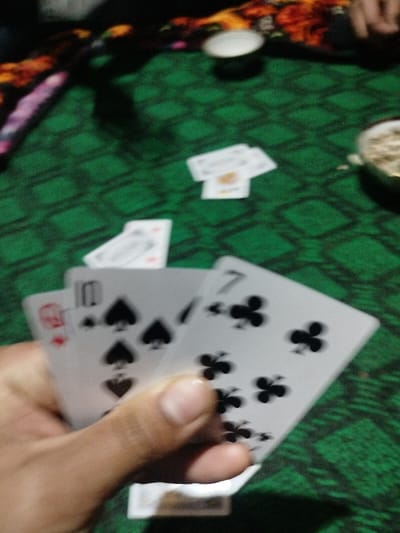 A hand holds three playing cards: an Ace of Hearts, a 10 of Spades, and a 7 of Clubs. Additional cards are scattered on a green patterned surface, along with a small white container and a bowl with contents suggesting snacks. The cards and containers are positioned on a table, with a decorative edge visible in the background.