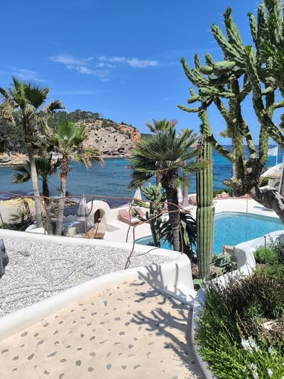 Captured in Sant Joan de Labritja, Spain, the image presents a serene scene with a blend of natural and constructed elements. Turquoise water stretches towards a rocky coastline under a clear blue sky dotted with sparse clouds. The foreground features a winding walkway with a light-colored, mottled surface, leading towards a terrace area. White architectural structures, including rounded walls and seating areas, are adorned with white pebbles. Palm trees and various cacti species, including a tall, columnar variety, punctuate the landscape. A rectangular swimming pool reflects the sunlight, and vegetation consisting of green plants and shrubs occupy the lower right corner, contributing to the overall Mediterranean ambiance.