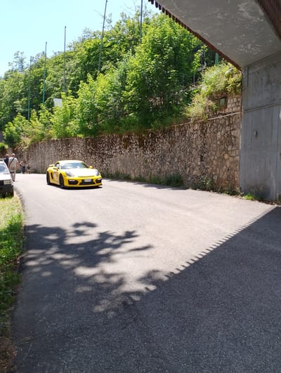 A bright yellow Porsche Cayman drives on a paved road with a low stone wall on the right and lush green trees on a hillside on the left. The car has a license plate that reads "EE 559XA". Two people are walking on the left side of the road, and a part of another car is visible on the far left. The sun casts shadows of trees and a fence onto the road. The background shows a clear blue sky and a concrete structure overhang on the right. This scene appears to be located in the city of Rieti, Italy, potentially on a scenic route or during an event that allows for such a display.