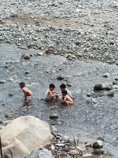 This image depicts five young children playing in a shallow, rocky river in Meurah, Indonesia. The water is clear, with visible ripples and reflections, and flows over a bed composed primarily of smooth, grey, and light-colored river stones of various sizes.
On the far left, one child, possibly nude, stands partially submerged in the water, facing towards the right. In the center, two children squat in the water; one wears a white tank top and holds a green plastic container, while the other, wearing light-colored underwear, looks towards the container. Further to the right, two additional children are partially visible, also squatting in the water, engaged in similar play.
The riverbanks are densely covered with similar river stones and pebbles, with occasional patches of exposed mud or sand. Scattered organic debris, such as dry leaves, is visible among the stones on the banks. Evidence of human litter includes a light blue plastic item partially submerged near the left child and a broken brown earthenware pot or jug resting on the rocky bank in the upper right quadrant of the image. The scene is illuminated by ambient daylight, suggesting an outdoor environment.