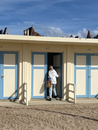 The image depicts a row of beach huts in Trouville-sur-Mer, France, under a bright blue sky with scattered clouds. The huts are painted a pale yellow with light blue trim around the doors and windows. Numbers are painted on the exterior of the huts, with "38" visible on the left, "67" on the middle hut, and "66" on the right hut. A person, viewed from the back, is entering the middle hut (number 67). This individual is wearing a light-colored jacket, blue jeans, and dark shoes. They are carrying a woven straw bag in their left hand. The ground in front of the huts is sandy beach, with footprints visible in the sand. In the background, the rooftops of buildings in Trouville-sur-Mer can be seen, characterized by steep gabled roofs and chimneys, suggesting an urban coastal environment. The lighting suggests it is daytime, possibly morning or afternoon, with clear shadows cast by the huts and a railing in front of them.