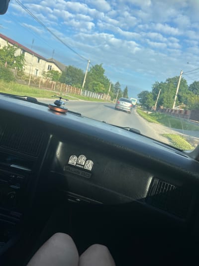 The image provides an interior view from a car driving on a sunny day in Velyki Hai, Ukraine. The dashboard features an "Audi 80" emblem and a small helicopter-shaped air freshener. Outside the car, a paved road is visible with houses and trees lining the street. A silver car is driving ahead on the road. The sky is filled with fluffy white clouds against a clear blue backdrop, suggesting pleasant weather. The time of day appears to be afternoon, given the bright sunlight and the shadows cast. The overall scene depicts a peaceful drive through a residential area.