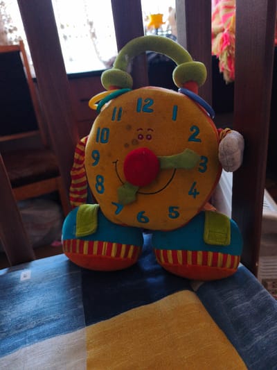 A stuffed toy clock with a yellow face, blue numbers, red nose, green hands, and a green arch on top is sitting on a chair in Nerubaiske, Ukraine. The toy is positioned in the center of the image, facing forward. The chair it is sitting on has a blue and yellow checkered cushion. In the background, wooden chair backs and a curtain are visible. The lighting suggests it is daytime, with light filtering through the curtain.