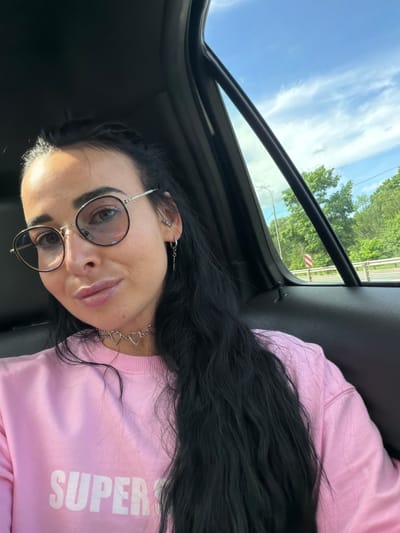 A woman with dark hair and glasses is in the backseat of a car. She is wearing a pink sweatshirt that says "SUPER" and a necklace with heart charms. Outside the car window, trees and a cloudy blue sky are visible. The photo was taken in the city of Mshaga, Russia.