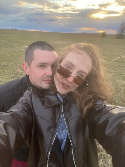 This image captures a couple, a man and a woman, posing closely for a photo outdoors in what appears to be a vast open field in Kueda, Russia, during either sunrise or sunset. The scene is illuminated by the "golden hour," with warm, golden light bursting through clouds on one side of the sky.
The woman, positioned slightly in front, has long, wind-blown, light brown hair. She is wearing stylish rectangular sunglasses with brown-tinted lenses, revealing glimpses of her light blue eyes. She sports a subtle smile and is dressed in a black leather jacket over a blue and white striped shirt.
The man is situated slightly behind and below her. He has short, dark hair and light stubble on his face, displaying a neutral to gently smiling expression. He is also wearing a black leather jacket.
The background showcases an expansive green field extending to a distant horizon dotted with sparse trees and possibly hills. The sky, a prominent feature, is filled with scattered clouds beautifully lit by the low sun. The overall atmosphere conveys a sense of affection and tranquility, capturing a shared moment between the couple in a picturesque natural setting. There is no visible text in the image.