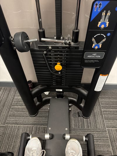 This is an image of a gym setting, likely in Watford, United Kingdom. The primary object in focus is a hip abduction machine, a piece of fitness equipment designed for exercising the hip muscles. The machine features a stack of weights, ranging from 10 lbs to 220 lbs (or 4.5 kg to 100 kg), with a selector pin visible for adjusting the resistance. Instructional diagrams on the machine illustrate how to perform the hip abduction exercise.
The scene is captured from a slightly elevated perspective, looking down at the machine and a portion of the surrounding gym floor. The floor is covered with grey and white striped carpet tiles. One person's feet, wearing white athletic shoes, are visible, indicating they are either using the machine or are near it. The gym appears to be indoors, with artificial lighting and neutral-colored walls, typical of a commercial fitness facility. No specific time of day or weather conditions are discernible. The overall impression is a functional and clean gym environment. The visible text includes weight denominations in both pounds and kilograms, as well as instructions for operating the machine and a warning label. The brand name "TENZIN" is also visible on a component of the machine.