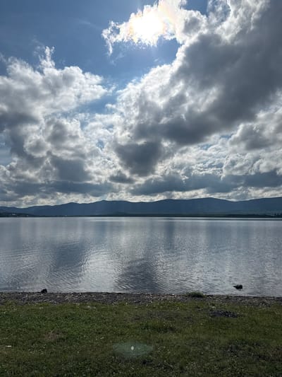 A wide shot captures a serene lake with calm, reflective waters. In the distance, a range of dark, silhouetted mountains forms the horizon. The sky is dramatic, with a mix of bright blue patches and large, billowing gray clouds. Sunlight pierces through the clouds, creating a bright halo and a subtle rainbow effect. The foreground features a narrow strip of rocky shoreline, littered with driftwood and debris, transitioning into a grassy bank. Two dark objects, possibly ducks or rocks, are visible in the water near the shore. The scene is devoid of any human presence. The overall impression is one of natural beauty and tranquility under a dynamic sky.