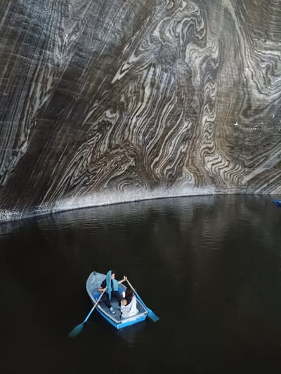 The image depicts an underground salt mine in Turda, Romania, specifically the Salina Turda. The foreground features a dark, still body of water, likely a lake formed within the mine. A small blue and white rowboat is in the water, carrying two individuals. One person, wearing a blue headscarf, is seated and holding oars, appearing to be rowing. The second person, wearing white, is seated in the back of the boat. The background is dominated by the massive, textured walls of the salt mine, characterized by dramatic striations of dark grey and white salt deposits. These geological formations create a surreal and cavernous atmosphere. The lighting is dim, highlighting the textures of the salt walls and the reflections on the water.