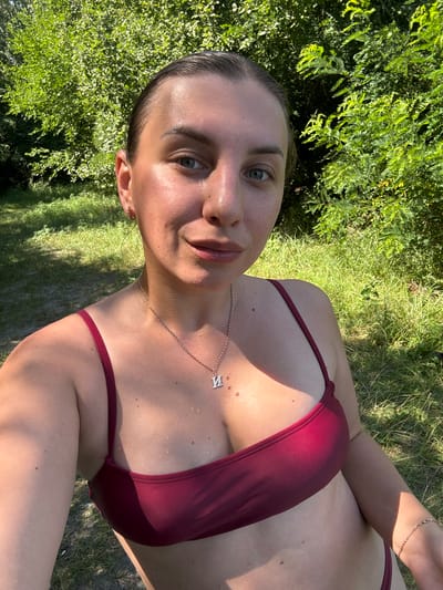 A woman is taking a selfie outdoors. She is wearing a burgundy bikini top and a silver necklace with a pendant. Her hair is pulled back, and she has a light-colored complexion. She is smiling slightly. The background is filled with lush green trees and grass, suggesting a natural outdoor setting, possibly a park or a wooded area. The lighting indicates it is daytime, with sunlight filtering through the foliage. The image appears to have been taken in Chykalivka, Ukraine, given the context.