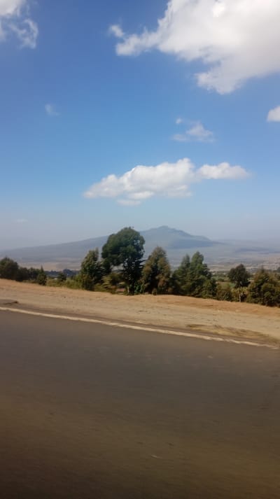 A scenic view features a road in the foreground, transitioning to a sandy shoulder. Beyond that, trees line the horizon with a distant mountain visible in the hazy background. The sky is a bright blue, dotted with scattered white clouds. The overall impression is that of a rural, elevated landscape under a clear sky.
