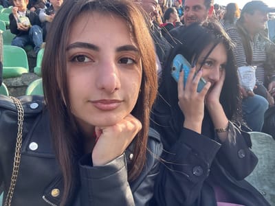 The scene is set in the spectator seating area of what appears to be a stadium or arena in Lanchkhuti, Georgia. The image captures two young adult females in the foreground, with a crowd of other individuals blurred in the background.
The female on the left has dark brown hair and is wearing a black leather jacket with silver button details and a visible silver chain from a shoulder bag. Her right hand is positioned under her chin, displaying red nail polish. She looks directly towards the camera with a neutral expression.
The female on the right has long, dark hair and is wearing a dark coat. She holds a light blue smartphone with a decorative case to her left ear, indicating she is engaged in a phone call. Her right hand partially covers the lower portion of her face. Her left hand shows black nail polish, and her right hand shows light purple nail polish. She is looking upwards and to the left, away from the camera.
Behind the two foreground subjects, rows of light green stadium-style seats are visible, occupied by numerous spectators. A young boy holding a snack bag is seated behind the left female, and an adult male with a beard is smiling behind the right female. The lighting suggests an outdoor, daytime setting with direct sunlight.