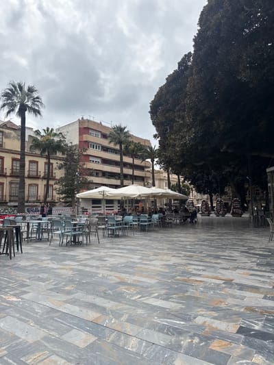 This image captures an outdoor urban plaza in Barriada De Villalba, Spain, under an overcast sky. The foreground is paved with rectangular tiles in varying shades of grey and light brown, arranged horizontally. In the mid-ground, a large open-air cafe features numerous light blue and grey tables and chairs, partially shaded by multiple white square umbrellas. A white kiosk, bearing "Mahou" branding, stands behind the seating area. Approximately 10-15 individuals are seated at the cafe tables, engaged in leisure activities. A red and white striped barrier tape extends across the left portion of the cafe area. To the left, two individuals walk past an older, multi-story building with a light-colored facade and reddish-brown window accents. The background features a taller, more modern multi-story building with a white and reddish-brown banded exterior, flanked by several tall palm trees. A very large, dense tree with dark foliage dominates the right side of the frame, partially obscuring a classically-styled building. Three tiered decorative planters, appearing to contain flowers, are arranged in a line on the right side of the plaza. The entire scene is illuminated by diffused natural light.
