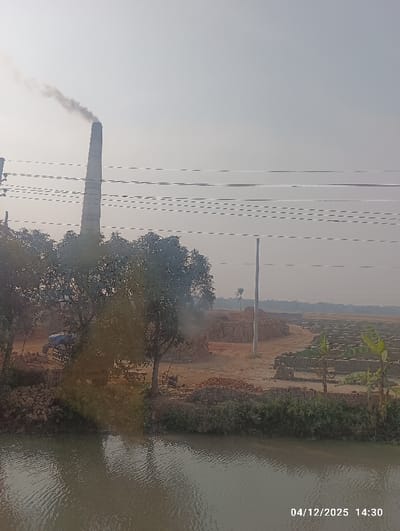 This image depicts an industrial landscape in Kaliganj, Bangladesh, at 14:30 on December 4, 2025. The scene is dominated by a tall, slender chimney, likely belonging to a brick kiln, positioned to the left of center, emitting a plume of dark grey smoke into a hazy, light-grey sky.
In the foreground, a body of water, possibly a canal or river, reflects the ambient light with rippling surfaces. Beyond the water, an unpaved, earthy expanse extends, filled with numerous neatly stacked rows and large piles of bricks, indicating a significant brick-making operation. Several green-leafed trees are scattered across the scene, particularly along the water's edge and near the base of the chimney. A blue tractor or similar vehicle is partially visible among the trees on the left. Overhead, multiple horizontal power lines or communication cables traverse the frame. A single white utility pole stands upright on the right side of the frame, reaching towards the cables. No individuals are visible within the frame. A single palm tree is distantly visible on the right horizon.