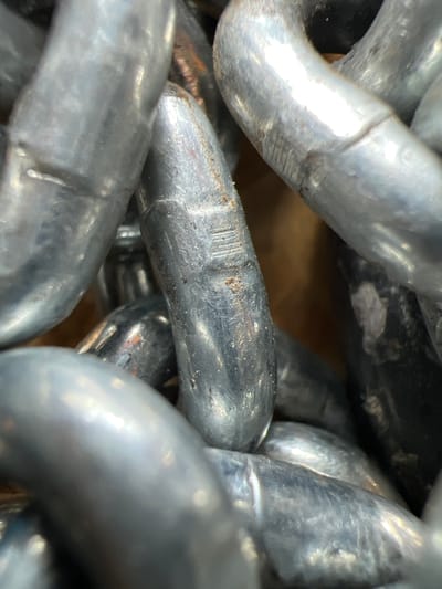 The image is a close-up, macro photograph of a pile of heavy-duty metal chains, likely made of steel. The chains are interlocking and appear to be of industrial or marine grade, with each link being thick and oval-shaped. The surface of the chains exhibits a metallic sheen, with areas of wear and some minor surface rust or discoloration visible, particularly on the central link which shows some fine horizontal lines or scoring. The lighting creates strong highlights and shadows, emphasizing the texture and volume of the metal. The background is out of focus, appearing as a warm, muted brown and black, suggesting the chains are stored in a dimly lit area. There are no people or any specific environmental context provided to pinpoint a location within Lanžhot, Czechia; the image focuses solely on the material and texture of the chains.