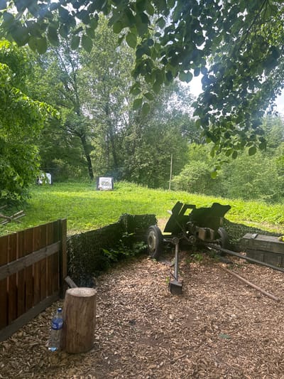 The image depicts an outdoor scene with elements that suggest a historical reenactment or training area. In the foreground, there is a wooden fence and a large tree stump with a bottle of water next to it. A military-style cannon is positioned behind a camouflage net, with ammunition crates visible to the right. The ground is covered in wood chips. In the background, a grassy field is dotted with trees, and two targets, possibly for archery or shooting practice, are visible. The overall environment is lush and green, indicating a natural setting. The presence of the cannon and targets suggests a location related to military history or activities. It is plausible this scene is located in Shlisselburg, Russia, a city known for its historical significance, particularly its fortress.