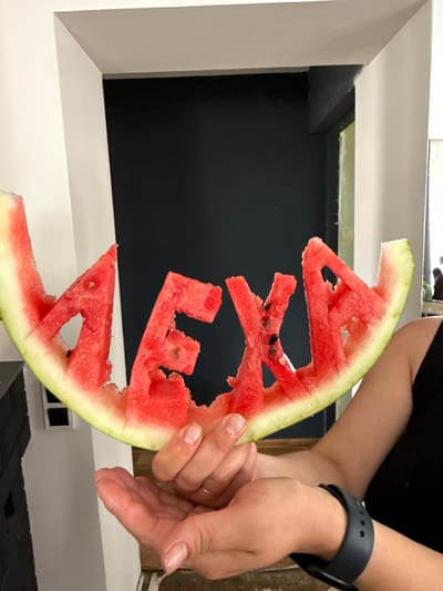 A person holds a slice of watermelon carved into the word "ALEXA" with their hands. The watermelon slice has a green rind and red flesh, with some black seeds visible. The letters are cut out from the flesh. The person is wearing a black top and a black watch on their left wrist. The background appears to be an interior space with a wall and a doorway. There is no information about the city of Хохряки, Russia in the image or its context.