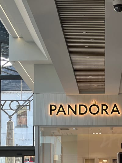The image captures the facade of a Pandora jewelry store, characterized by its illuminated sign featuring the brand name "PANDORA" in bold, black lettering with a warm, glowing outline. Above the text, a small, stylized crown symbol is also illuminated. The storefront features large glass windows, reflecting the interior and offering glimpses of display cases. The building's architecture includes modern design elements such as angled white panels above the sign and a slatted ceiling with recessed lighting. A security camera is visible mounted on the upper right corner of the ceiling. To the left of the Pandora store, through a glass window, parts of another establishment are visible, including a white building with the partial word "TOMMY" in block letters. The overall setting appears to be an indoor shopping environment, possibly a mall or a modern commercial street, with elements of structural design and retail branding. The scene is devoid of any individuals. The location is identified as the city of Derry/Londonderry, United Kingdom.