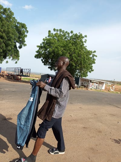 In Gadao, Nigeria, an elderly man with a bald head and a light beard walks on a dusty road. He is wearing a black and white striped long-sleeved shirt, dark trousers, dark socks, and black and white slide sandals. He is carrying a bundle of clothes in his left hand. The clothes include a blue denim garment with the letters "KK" printed on it, and another dark-colored garment. He is draped with a brown cloth over his shoulders and arms. The background shows a few trees, a large trailer, and a small roadside stall with goods displayed. The sky is partly cloudy with a light blue hue.