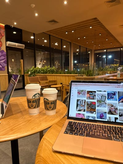 The image presents an interior view of a Starbucks cafe located in Santa Tecla, El Salvador, observed during nighttime. In the immediate foreground, two adjacent light-colored wooden round tables are visible. The table on the left hosts a silver/white laptop, partially open and standing upright, alongside two white Starbucks paper cups with brown logo sleeves, one larger than the other. The table on the right features an open rose-gold MacBook Air laptop, its screen displaying a grid of various lifestyle and aesthetic images, consistent with a social media platform like Pinterest.
The background reveals the cafe's internal layout, characterized by large, floor-to-ceiling glass windows that look out onto a dark exterior with visible distant lights. Inside, wooden slatted panels adorn sections of the walls and partitions. Green potted plants are strategically placed along the base of the windows and behind seating areas. Beyond the foreground tables, brown upholstered armchairs and small wooden tables are arranged. The ceiling is light-colored, equipped with multiple recessed light fixtures and decorative wooden slats. A security camera is mounted on the upper left wall, adjacent to a horizontal air conditioning unit. A colorful, abstract mural, featuring floral or plant motifs, is visible on the far-left wall. No individuals are depicted within the scene.