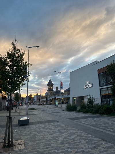 The image captures a street scene in Eastbourne, United Kingdom, at dusk or dawn. The sky is filled with clouds, illuminated by a soft, golden light. Dominating the mid-ground is the Eastbourne railway station clock tower. To the left is a modern building with the inscription "The Beacon" on its facade. The street is lined with lampposts and trees. The pavement is made of patterned blocks. A bus stop can be seen. Cars are visible on the road in the distance. People are present but appear small and indistinct.