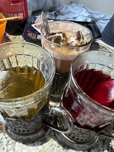 The image shows three glasses on a granite-patterned surface. In the foreground, two glasses are in ornate metal holders. The left glass contains a pale yellow liquid, likely tea. The right glass contains a dark red liquid, possibly compote or berry juice, with a few pinkish-red pieces floating within. The third glass, positioned behind and between the other two, contains a brown beverage topped with whipped cream and chocolate pieces. A silver spoon rests in this glass. To the left, partially visible, is an orange container and a red bottle with Cyrillic lettering, suggesting it might be a beverage. The background is blurred, showing fabric and other indistinct objects. The overall scene suggests a casual setting where beverages are being consumed. The mention of Gorodische, Russia, implies this image was taken in that city.