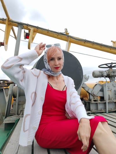 A woman with pink hair, wearing a red dress and a white cardigan with a heart pattern, sits on a wooden deck. She has a patterned scarf tied around her head and is holding her sunglasses on top of her head. She is looking at the camera with a neutral expression. Behind her, there is industrial equipment, including a large black circular object, some yellow metal structures, and grey machinery with a large steering wheel. A red light is visible on one of the yellow structures. The sky is overcast, suggesting cloudy weather. The scene appears to be on a boat or a dock. No visible text is present. The location context is Novokemskii, Russia.
