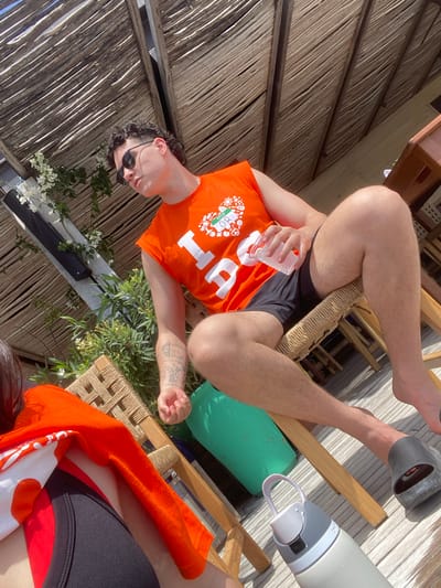 The image shows a man wearing an orange "HELLO OTTAWA" shirt, black shorts, and sunglasses. He is sitting on a woven chair, holding a glass of water. A water bottle and a green cushion are next to him. He has a tattoo on his left arm. In the background, there is a thatched roof, suggesting a beach or resort setting. White flowers and green plants are also visible. The location is Marina Maroma, Mexico.