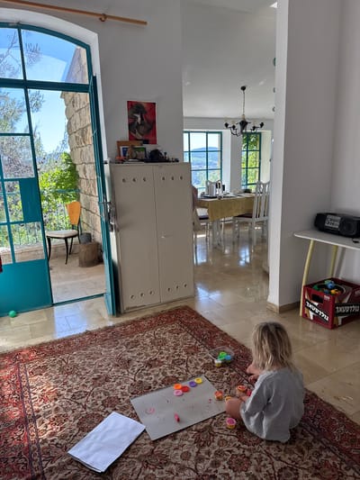 A bright, sunlit indoor scene captures a child engrossed in creative play within a home environment. A young child with light hair, wearing a grey long-sleeved top, is seated on a large, ornate red and beige patterned rug. They are focused on an activity involving colorful small containers, possibly paint or play-doh, arranged on a grey drawing pad that shows smudges of color. A folded white paper rests nearby on the rug.
The room features light-colored tiled flooring and white walls. Large windows with distinctive green frames allow ample natural light into the space and offer views of lush outdoor greenery, trees, and stone architecture, suggesting a sunny day. One window on the left has an arched top.
In the midground, a large white metal cabinet with circular details stands against a wall. Further into the background, a dining area is visible with a table, light tablecloth, and several white chairs, illuminated by a black chandelier. Above a section of the wall, a black boombox with an "ICC" logo rests on a white shelf, next to a red cardboard box. This box has Hebrew text, "גולדטאר" (Goldstar), and "500" printed on it, along with "GOLDASTAR" and "MADE IN CHINA". A picture depicting two figures, possibly animals, hangs on a wall near the cabinet. A small green ball is on the tiled floor near a doorway leading outdoors, where a wooden stool or chair is visible. The bright natural light indicates it is daytime.