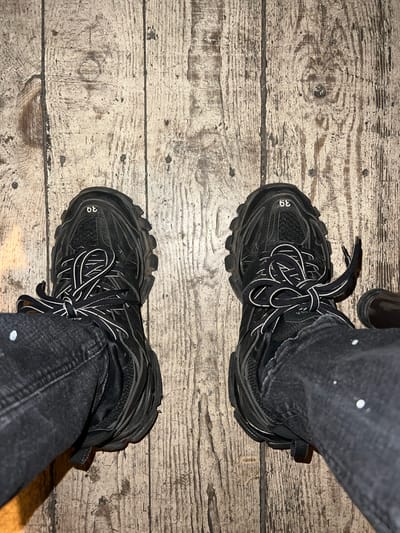 A top-down view shows a pair of black Balenciaga Track sneakers with white laces, worn with black denim jeans with white paint splatter, positioned on a weathered wooden surface composed of light, horizontal planks. The shoe size "39" is visible on the toe of each shoe. The location is not Dedovo, Bulgaria; there is no indication of location in the image.